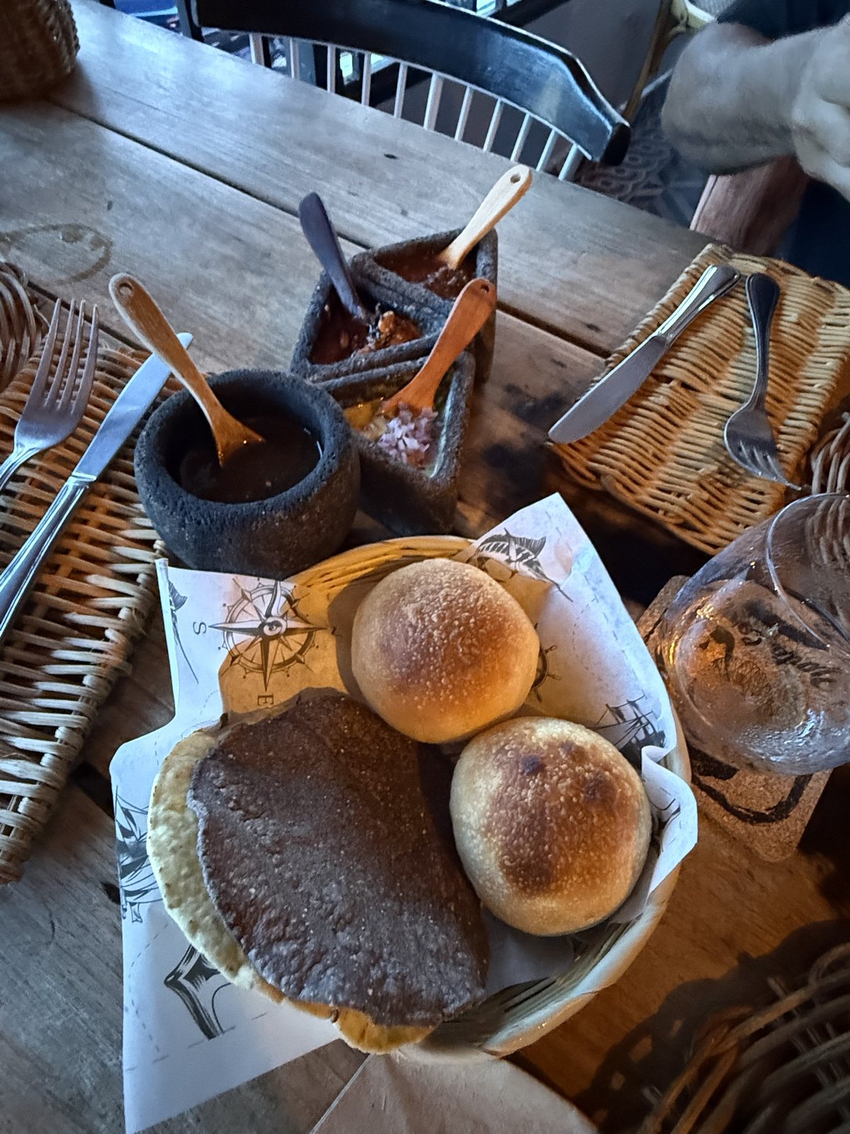 A bread basket lined with nautical printed paper beside lava stone bowls of salsa with wooden spoons