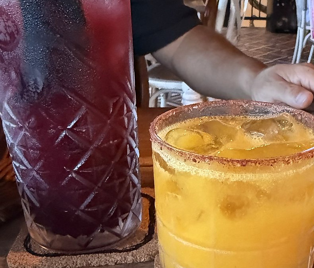 Two cocktails side by side on a wood table: a dark red Rojito in a cut glass and a yellow Tepache Jam with a chile salt rim