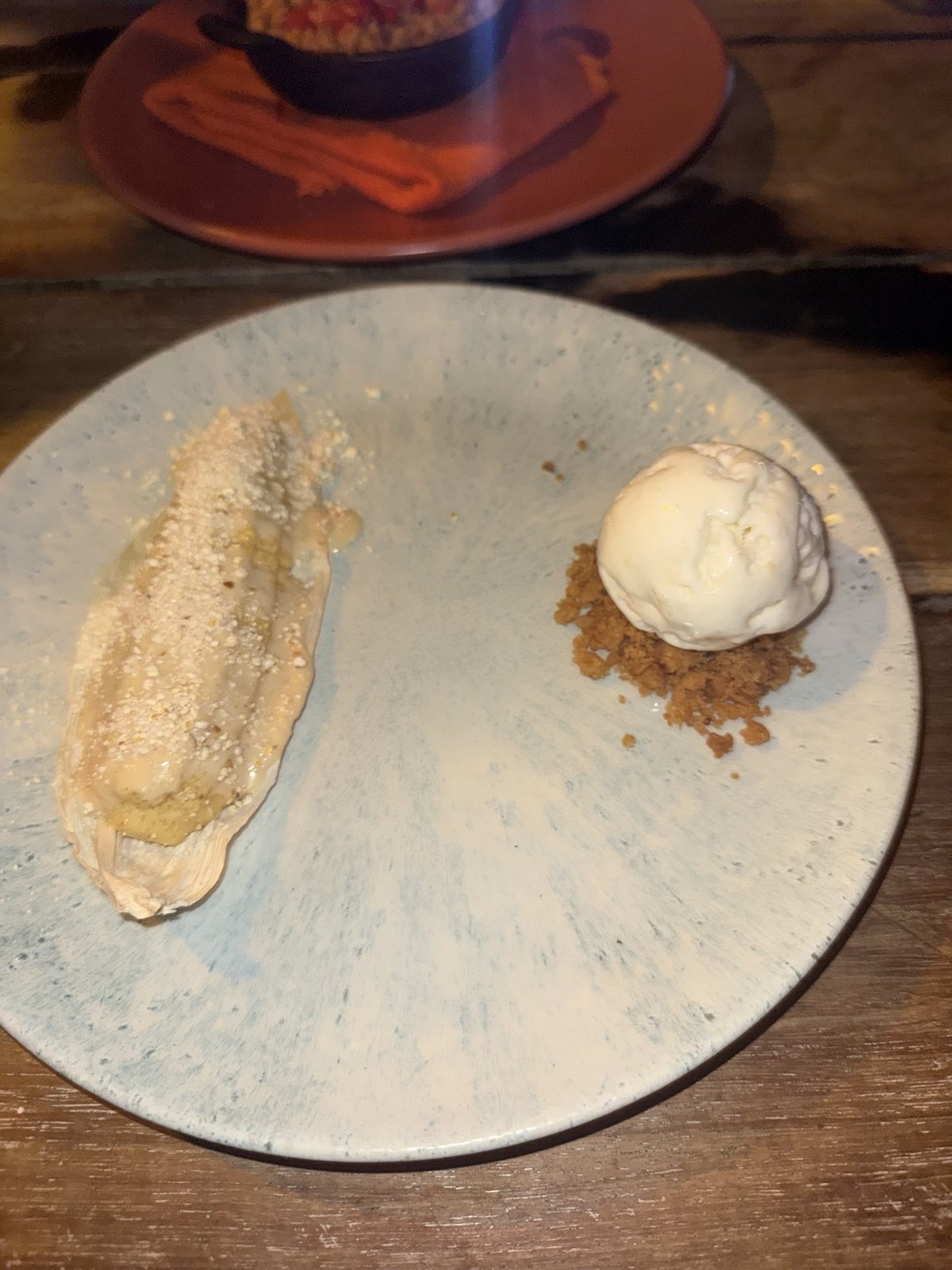 A sweet corn dessert served in a corn husk beside a scoop of ice cream and crumble on a pale blue ceramic plate