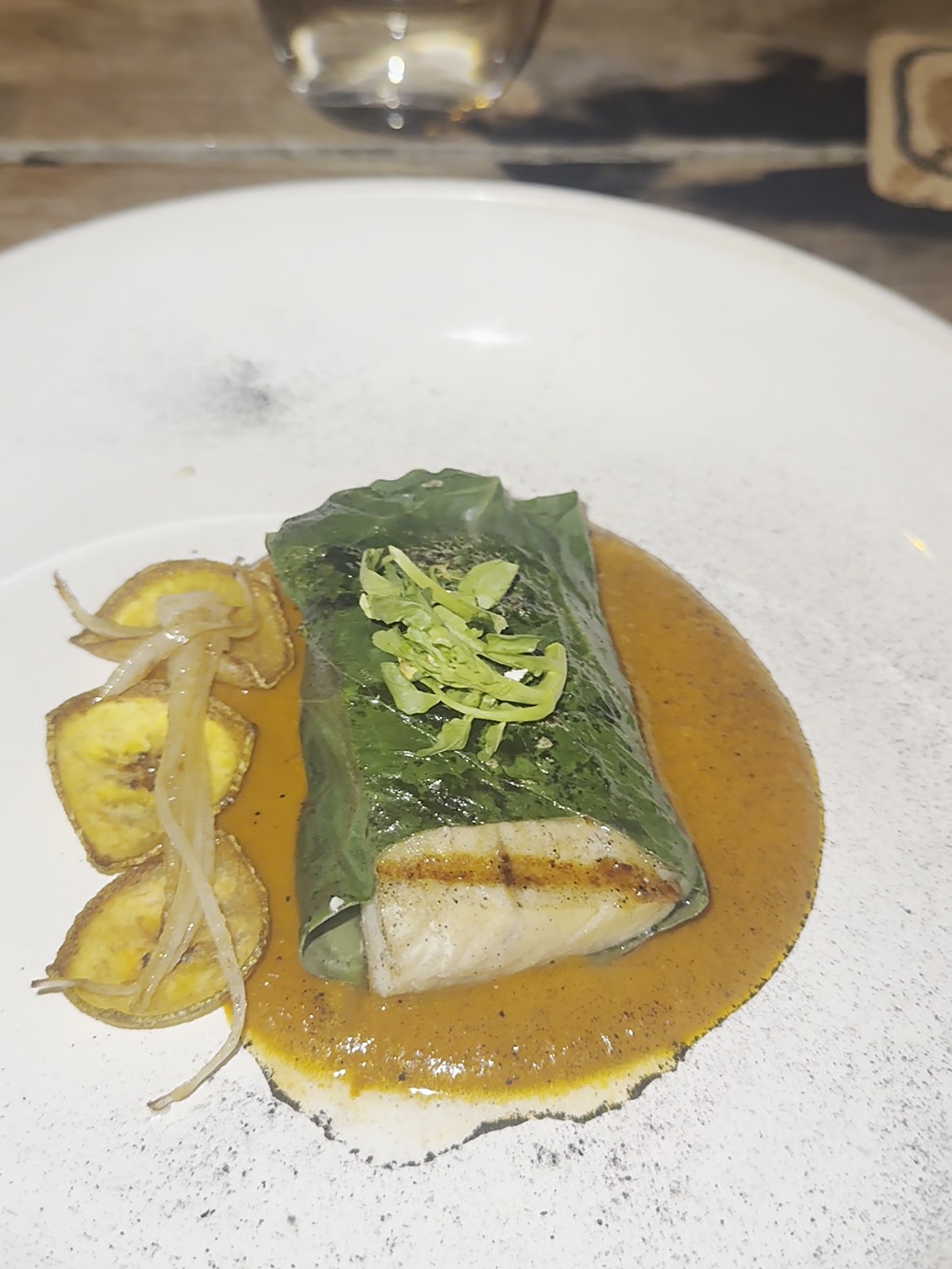 A fish fillet wrapped in Mexican pepper leaf on yellow mole, with fried plantain and grilled onions