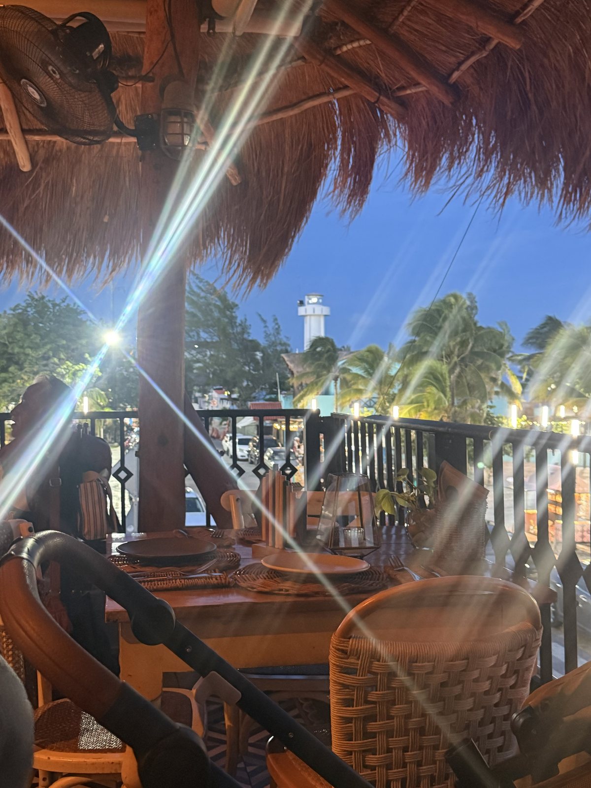 View from the restaurant at blue hour through a thatched roof toward the Puerto Morelos lighthouse and palm trees