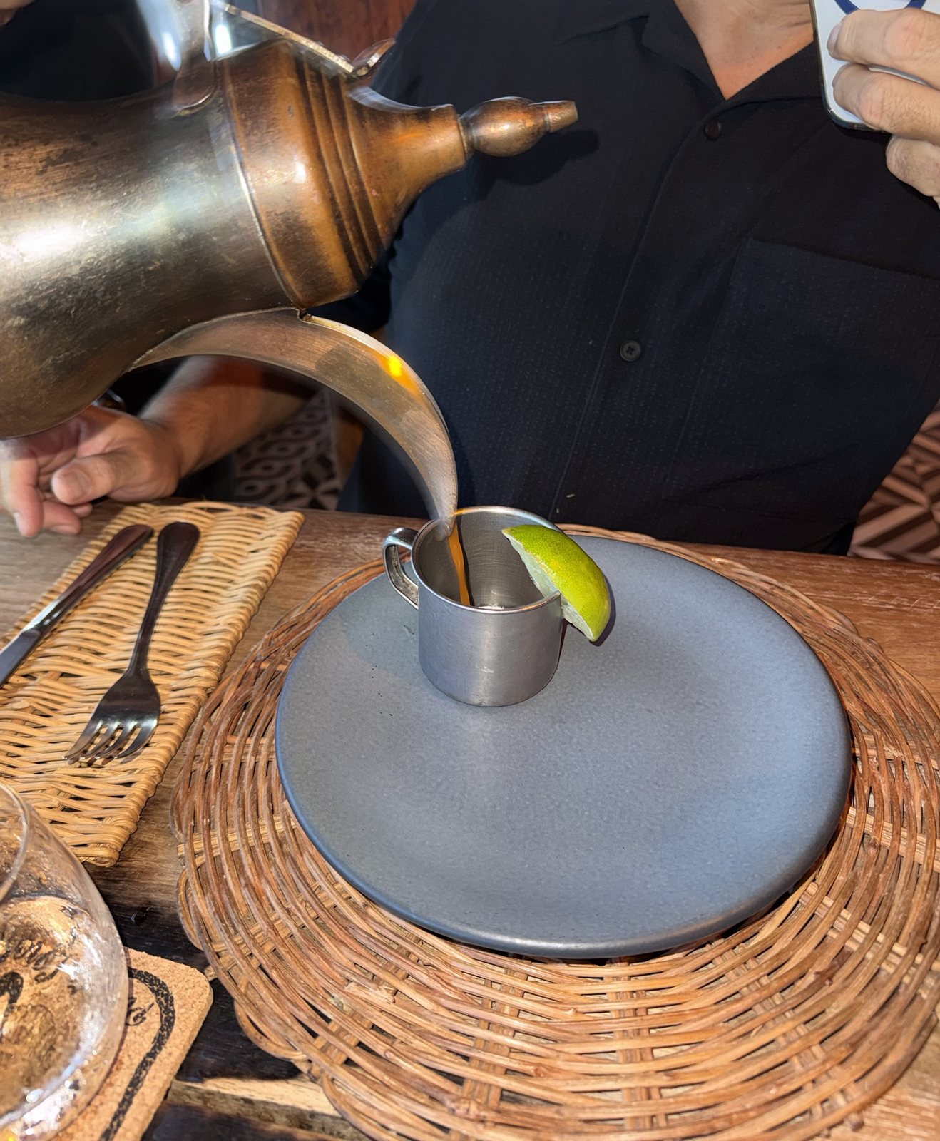 Shrimp soup being poured from a brass vessel into a small tin cup with a wedge of lime, on a dark plate
