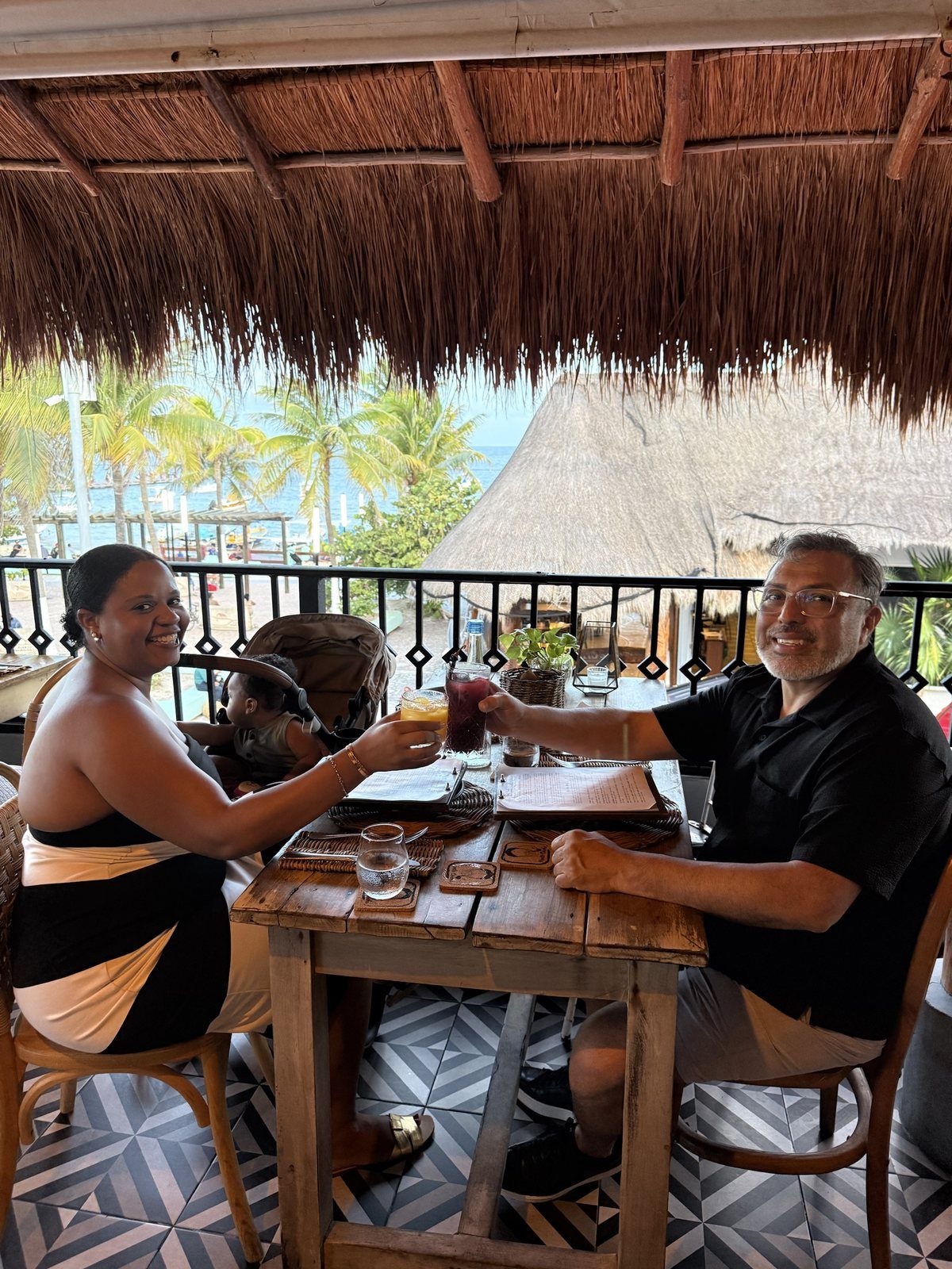 Two diners raising cocktails across a rustic wood table under a thatched roof, with palms and the Caribbean behind them